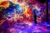 A man standing looking up at a bright purple, orange, and pink projection of the galaxy and stars on the walls and floor at ARTECHOUSE NYC.