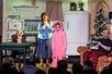 A young boy with glasses in a pink bunny suit with his mother, father, and brother around him in a living room on stage at the A Christmas Story Dinner Show.
