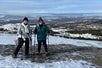 Two guests wearing cold-weather hiking attire and snowshoes and holding trekking poles as they smile at the camera with South Fork Campbell Creek valley in the background.
