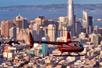 A red helicopter flying through the air with the city of San Francisco in the background at sun set on the Alcatraz and San Francisco Sites Helicopter Tour.