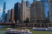Several tour boats full of people on the Architecture River with the city of Chicago on the other side on a sunny day.