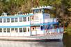 The front of the Barefoot Queen Riverboat Cruise in the water with the sun shining on it and trees in the background.