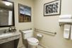 A private bathroom with a small sink and mirror on the left with a toilet next to it plus grab bars, a towel rack, and art on the walls.