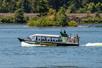 Wide shot of the profile of Explorer Jetboat will of tourists in motion on a sunny day with trees in the background.