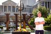 Gordan Ramsay in a white chefs jacket standing near the on fire sign for Hell's Kitchen on a sunny day with Caesar's Palace in the background.