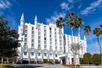View of the front of the Castle Hotel, a large white hotel with spires at the top and palm trees in front of it on a sunny day in Orlando.