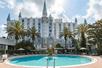An outdoor swimming pool with a fountain in the middle and lounge chairs and large palm trees around it with the Castle Hotel in Orlando, Florida.