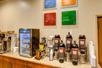 A coffee station with several different types of coffee in dispensers with cups to the right and a mini fridge with other drink options to the left.