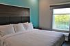 A hotel room with a white king bed, blue accent wall, and mirror with the blinds open.