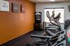 A fitness center with a row of  cardio equipment, free weights, a drink station, and an orange accent wall with art on it.