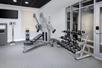 A fitness center with gray walls and floor and a weights area with a large bench and a free weights rack next to a window.