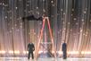 Criss Angel walking down the side of a ladder while being parallel to the floor with two men in black suits standing and watching him.