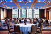 Conference/Banquet hall with round tables, chairs, large windows and carpeted floors.