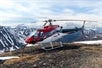 A red and white helicopter on the ground with a snowy mountain backdrop and blue skies peeping through the clouds
