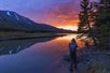 A photographer wearing a hoodie and backpack looks out onto a mountain lake reflecting the sunset and mountain backdrop