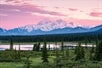 A mountain lake in Denali, Alaska set amidst a forest with a snow-capped mountain background and sky of pink