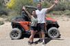 A woman in a green tank top and a man in a white shirt smiling with their arms up in front of a red ATV in the desert.