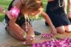 A woman in a black outfit kneeling on a woven rug stringing flowers together to make a lei at Diamond Head Luau.