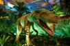 A dinosaur statue with with its mouth open standing a grassy area with several fake plants and in a low lit room decorated like a jungle.