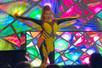 A drag queen wearing a skin tight yellow and black body suit with her arms out and a stained glass background behind her.