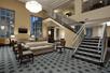 A large lobby with geometric carpet, several seating areas with beige and wood furniture, large windows, and stairs leading to the second floor.