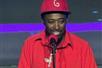 Eddie Griffin in a red track suit with a silver chair and a red hat performing in front of a microphone on stage.