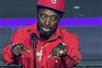 Eddie Griffin in a red track suit with a silver chair and a red hat performing with both hands up in front of a microphone on stage.