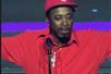 Eddie Griffin in a red track suit with a silver chair and a red hat performing with his arms out in front of a microphone on stage.