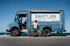 A man standing with a teal bicycle and leaning against te Drifters Beach and Bike Rentals blue delivery truck.