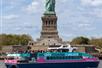 A teal and pink cruise boat with the top deck full of people sight seeing with the statue of liberty behind them.