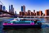 A teal and pink cruise boat with the top deck stuffed with people with the city in the background and the Brooklyn bridge to the left.
