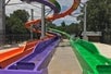 A colorful water slide attraction featuring multiple twisting slides in vibrant hues, with a clear view of the water channels below and a backdrop of green trees.