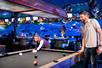 A man and woman playing a game of billiards on a black table with dining tables and a bowling alley in the background.