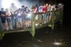 A large group of people leaning against a wooden railing and point flash lights into the alligator filled water below them.