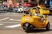 View of a yellow GoCar from behind on the street with two tourists wearing helmets driving it and a line of cars in front of it.