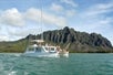 A boat with people on it sailing in the water as part of a Voyaging Catamaran Tour at Kualoa Ranch with cloudy skies a beautiful mountain in the background.