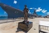 A bronze statue of Admiral William Halsey Jr. adorned with a lei, positioned on the dock in front of the USS Missouri, with clear blue skies and the battleship's hull visible in the background.