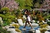 A woman steps on a stone in the water with cherry blossom and green trees in the background on a hot day at the Japanese Friendship Garden in Balboa Park.