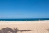 An empty beach with light colored sand and deep blue ocean water in the background on a sunny day.