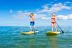 A man and woman in swim suits standing on yellow paddle boards in the ocean on a sunny day with clouds in the sky.