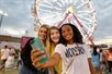 Three girls take a picture in front of the ferris wheel at the GrapeFest Carnival