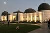 Tourists standing and sitting in front of the front of Griffith Observatory at dusk with the moon shining over head.