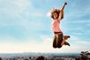 A woman with very curly hair wearing a pink tank top jumping in the air with her arms up on sunny day.