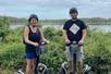 A man and woman wearing helmets and standing on segways with bushes and water behind them on the Guided Segway Tour of Lake Louisa State Park.