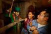 A leprechaun in a green suit with a bloody mouth and a club scaring a man and a woman who are both screaming.