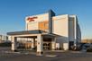 The front exterior and covered drive way and entrance of the Hampton Inn Portland Airport with the sun shining on it and clear sky over it.