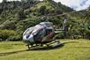 A silver and black Maverick helicopter parked in some grass in the Hana Rainforest with a mountain in the background on a sunny day.