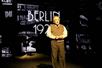 A short older man in khakis and a sweater vest with a white shirt under it standing in front of a wall that says "Berlin 1927" in the musical Harmony.