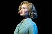 A woman in a light blue top with shorter blonde hair that is curled at the end with a sorrowful look on her face in the musical Harmony.