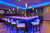 A "U" shaped bar with white counter tops and chairs and purple lights shining down on them plus a wall of liquor.